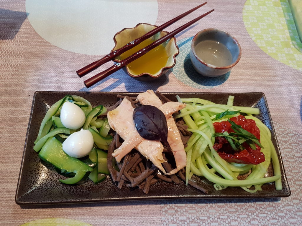 legumes nouilles soba et garniture sur une assiette marron rectangulaire avec une coupelle sauce et un verre de sake baguettes posees sur la coupelle de sauce