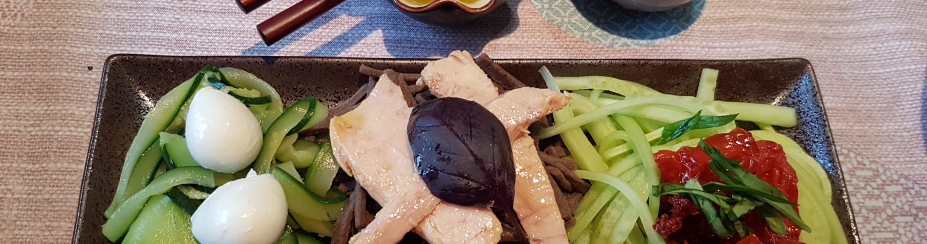 legumes nouilles soba et garniture sur une assiette marron rectangulaire avec une coupelle sauce et un verre de sake baguettes posees sur la coupelle de sauce