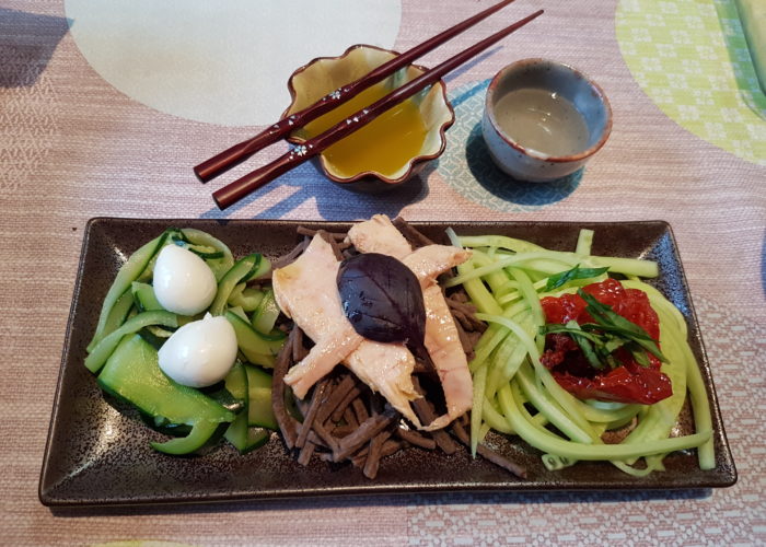 legumes nouilles soba et garniture sur une assiette marron rectangulaire avec une coupelle sauce et un verre de sake baguettes posees sur la coupelle de sauce