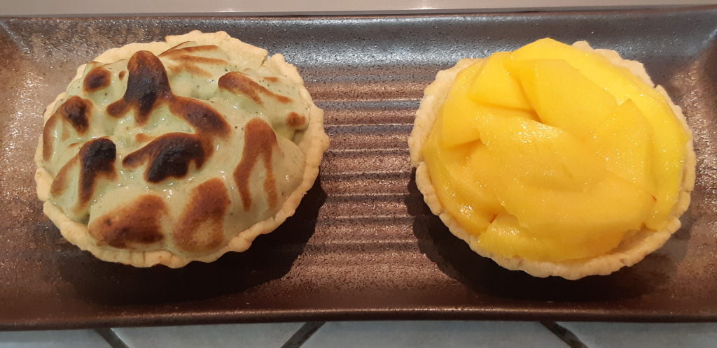 deux tartelettes sur une assiette rectangulaire sombre une avec une meringue verte legerement brulee, l'autre avec des tranches de mangue