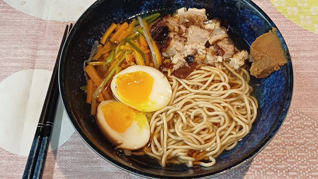 bol de ramen avec de la pâte miso su le bord droit on voit dans du bouillon clair des nouilles deux moities d'oeuf mollet du porc braisé et des legumes en julienne