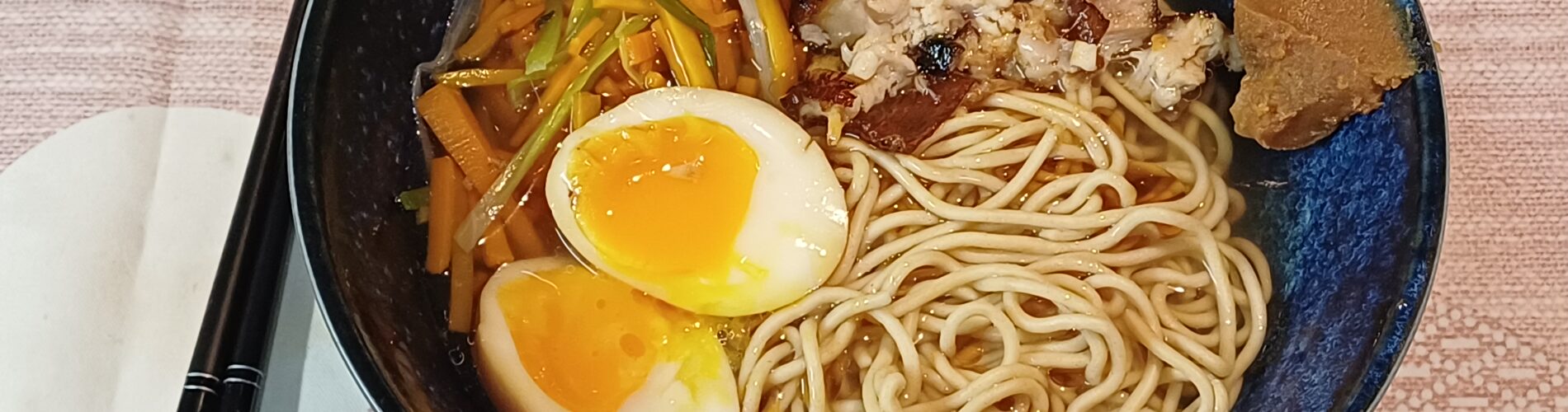 bol de ramen avec de la pâte miso su le bord droit on voit dans du bouillon clair des nouilles deux moities d'oeuf mollet du porc braisé et des legumes en julienne